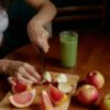 Person slicing fruits with a knife, next to a green juice and medicine on a wooden board.